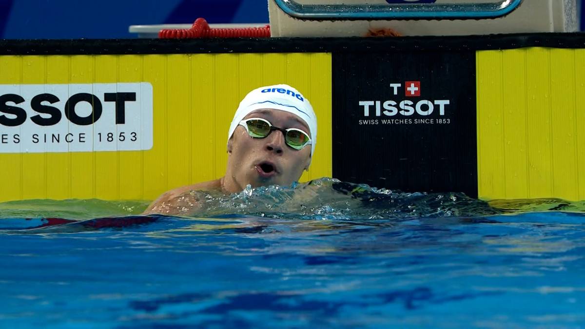 Pływak w czepku i okularach pływackich wykonuje ruch ramionami w basenie, z ustami uchylonymi do wydechu. W tle widoczna żółta i czarna tablica z logo Tissot.