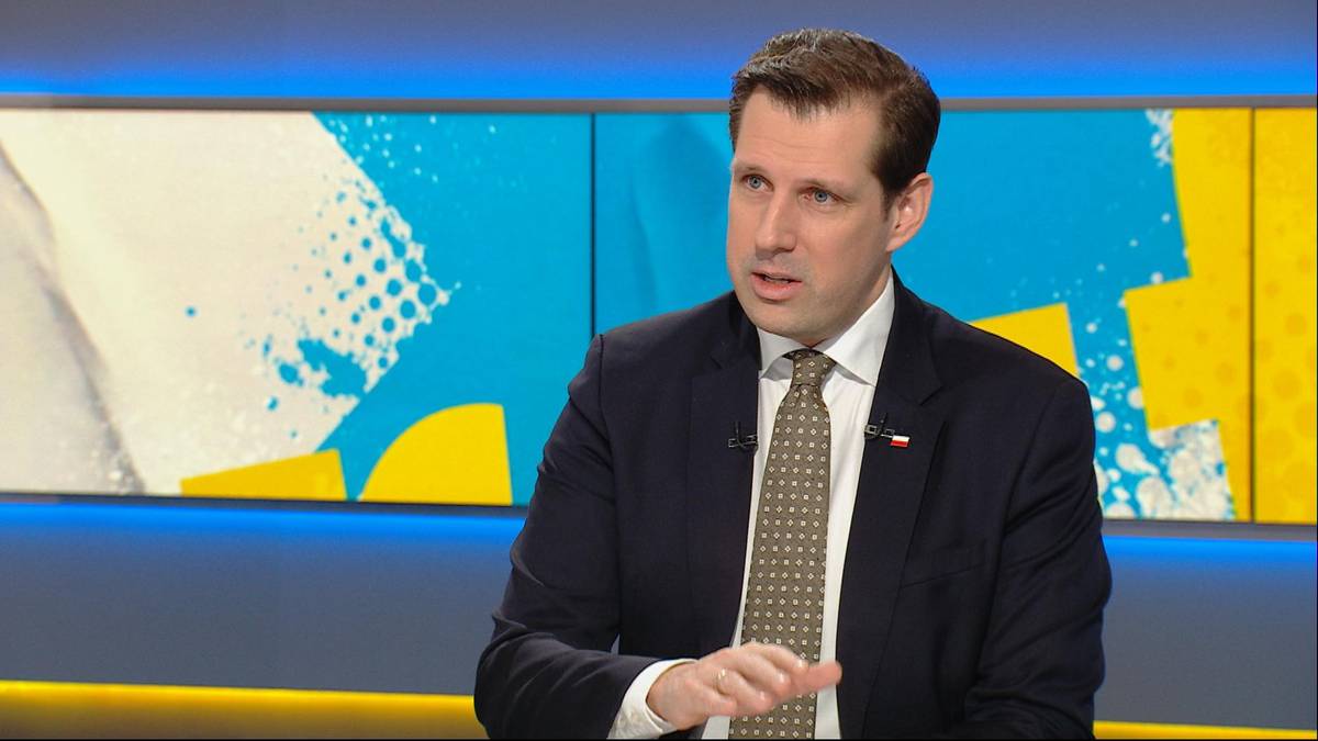 Mężczyzna w garniturze i krawacie siedzi w studiu telewizyjnym i gestykuluje.