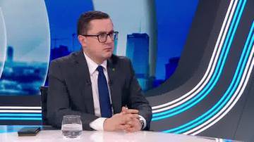 Mężczyzna w garniturze i okularach siedzi przy stole, rozmawia.