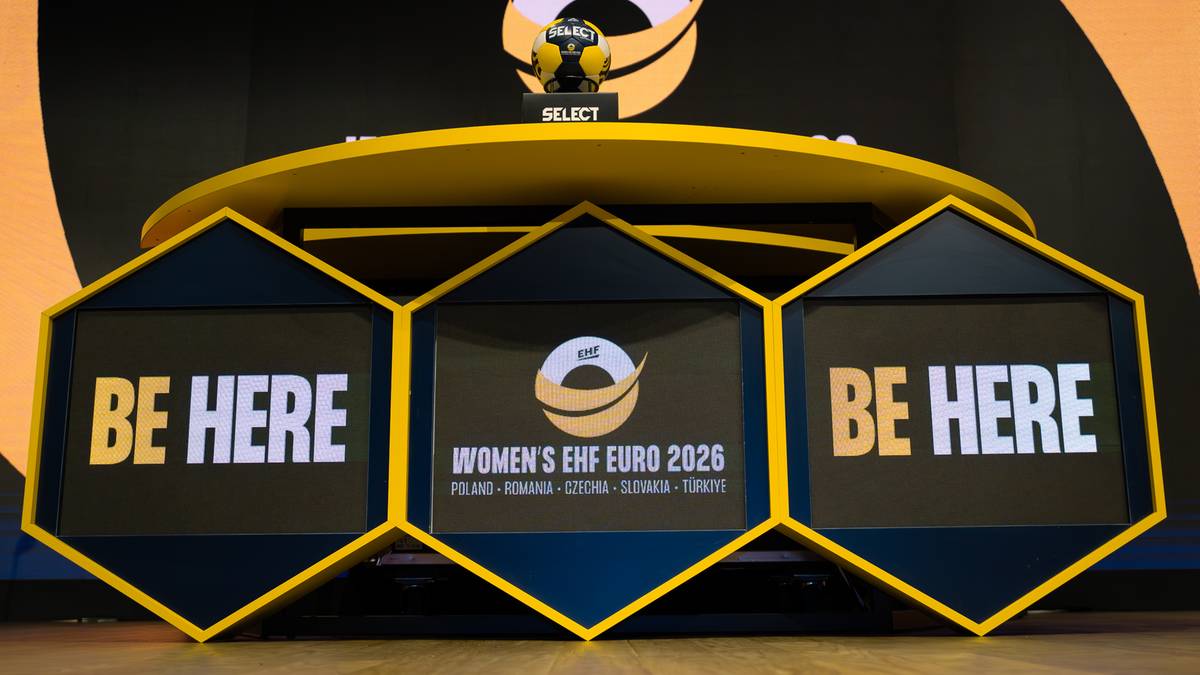 Stół z trzema hexagonalnymi ekranami. Na środkowym ekranie logo "WOMEN'S EHF EURO 2026". Na bocznych ekranach napis "BE HERE".