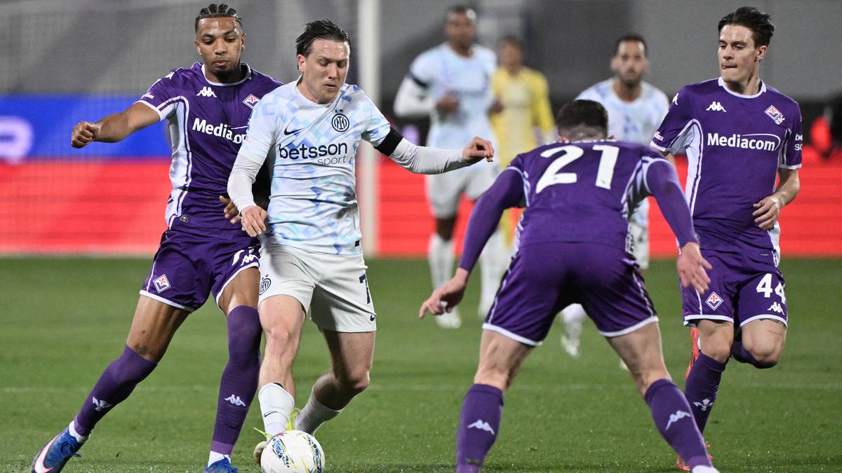 Piłkarze walczący o piłkę na boisku, jeden z nich w jasnym stroju, pozostali w fioletowych.