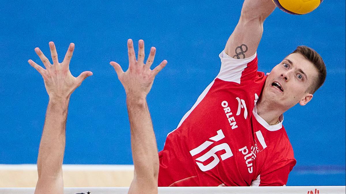 Gracz reprezentacji Polski w siatkówce w czerwonej koszulce z numerem 15 atakuje piłkę nad siatką. Obok widoczne dłonie innego zawodnika blokującego.