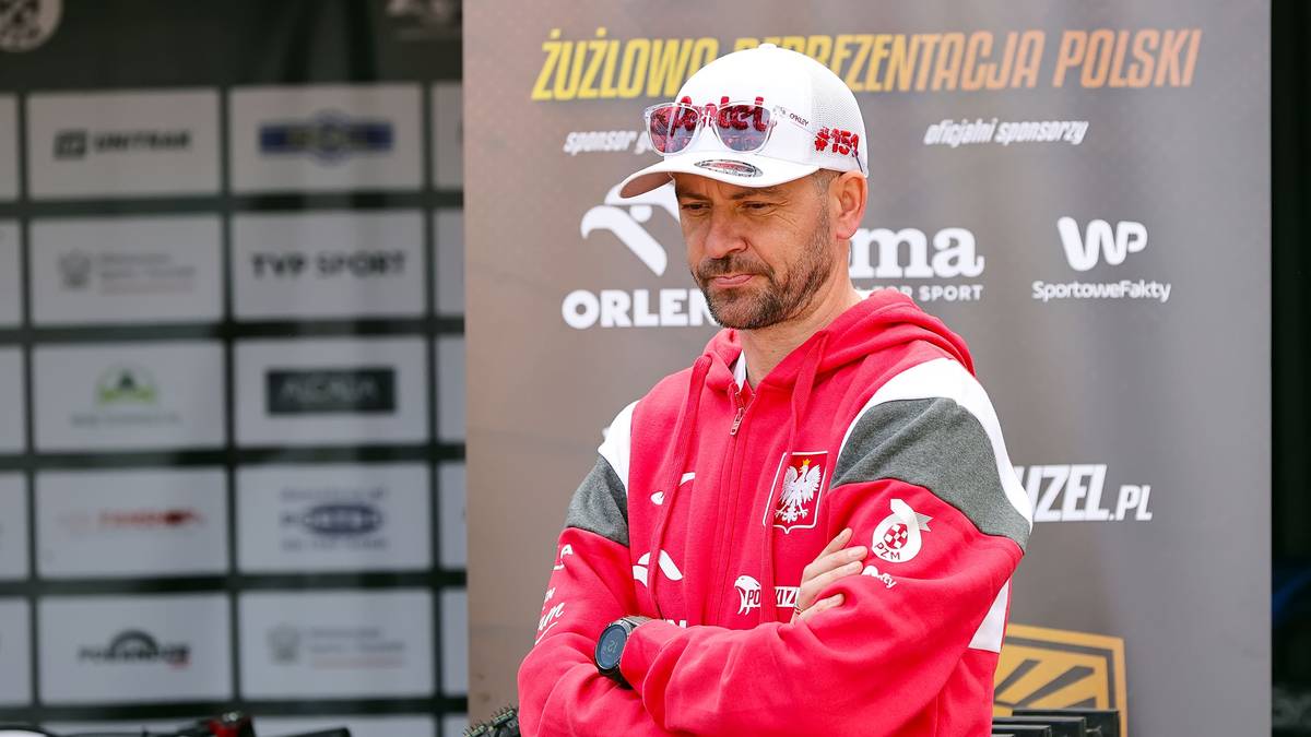 Mężczyzna w czapce z daszkiem i czerwonej bluzie z kapturem z herbem Polski, stoi ze skrzyżowanymi ramionami.