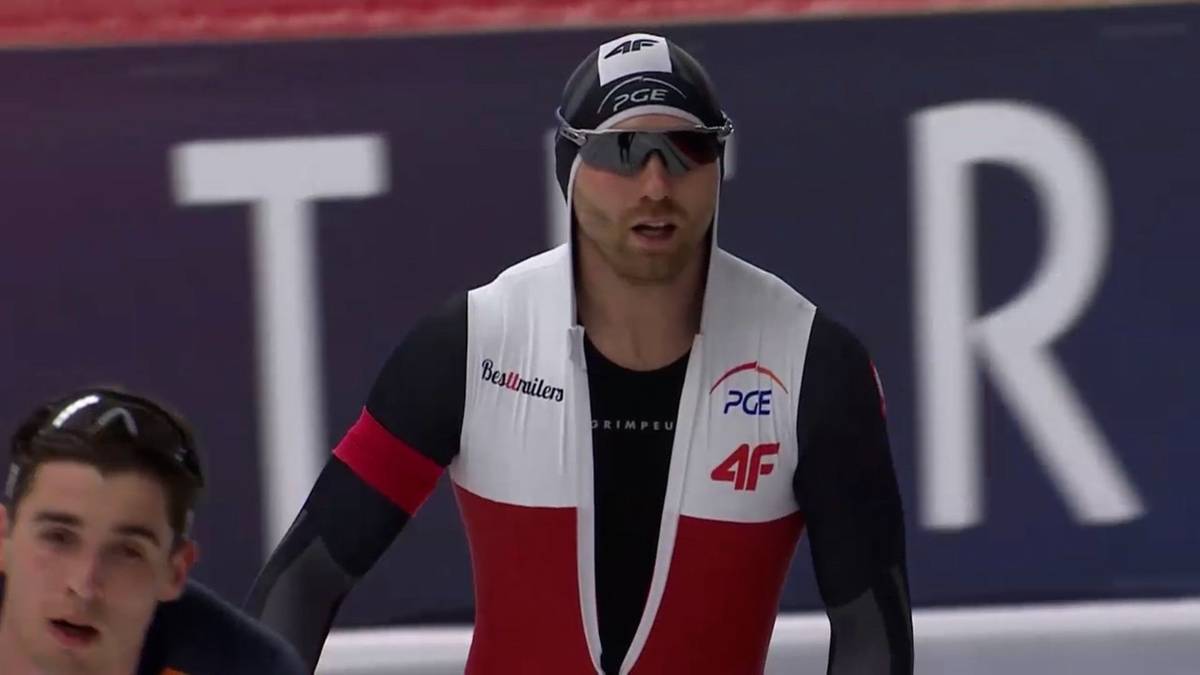 Łyżwiarz szybki w stroju sportowym z logo PCE i 4F, w goglach i czapce, przygotowuje się do startu.