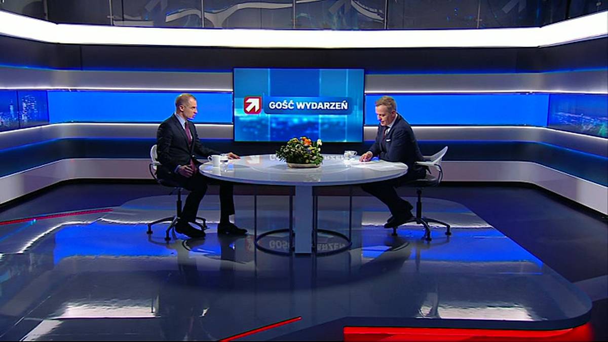 Dw&oacute;ch mężczyzn w garniturach siedzi przy okrągłym stole w studiu telewizyjnym.