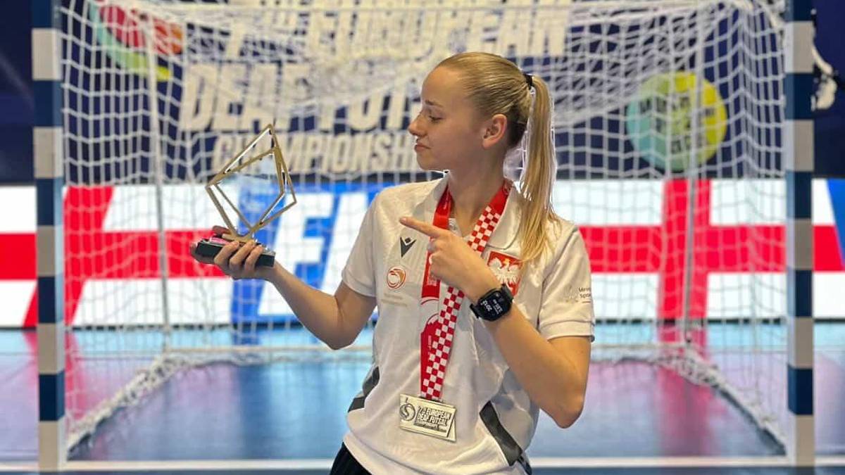 Zdjęcie młodej sportsmenki z pucharem i medalem w ręku, na tle bramki do piłki ręcznej.