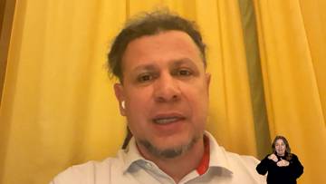 Mężczyzna z kilkudniowym zarostem i słuchawkami w uszach, ubrany w białą koszulkę polo z czerwonym kołnierzykiem, wypowiada się do kamery. W prawym dolnym rogu obrazu widoczna jest kobieta migająca w języku.