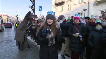 Grupa ludzi w koronasach na głowach, w tym młoda kobieta z mikrofonem, uczestnicząca w Orszaku Trzech Kr&oacute;li. Obok niej stoi osoba w przebraniu przypominającym smoka lub dinozaura.