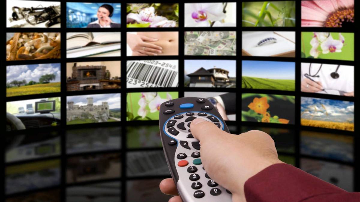 Dłoń trzymająca pilot do telewizora na tle wielu ekran&oacute;w telewizyjnych.