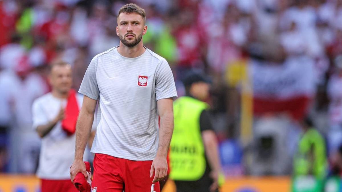 Piłkarz w szarym t-shircie z godłem Polski i czerwonych spodenkach, idący po boisku, na tle trybun wypełnionych kibicami w biało-czerwonych barwach.