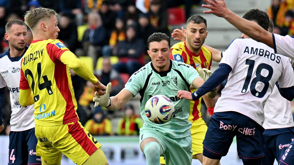 Piłkarze walczą o piłkę podczas meczu piłki nożnej. Bramkarz w zielonym stroju próbuje przejąć futbolówkę od zawodnika w żółto-czerwonej koszulce.