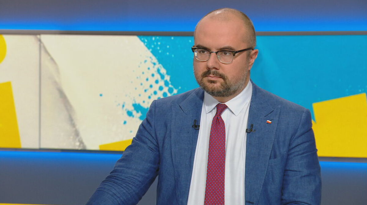 Mężczyzna w okularach, z zarostem, ubrany w marynarkę i krawat, siedzi przed ekranem z żółto-niebieskimi elementami graficznymi.