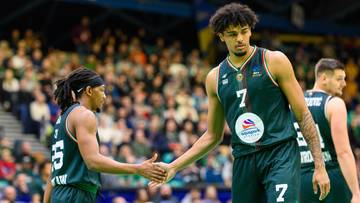 Dwóch koszykarzy w zielonych strojach ściska sobie dłonie na boisku, za nimi widać publiczność na trybunach.