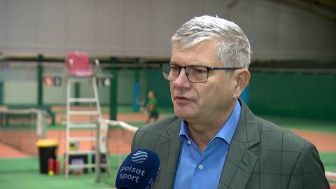 Zbliżenie na twarz Dariusza Łukaszewskiego, starszego mężczyzny w okularach, w niebieskiej koszuli i marynarce, m&oacute;wiącego do mikrofonu Polsat Sport. W tle kort tenisowy z widocznymi innymi osobami i sprzętem.