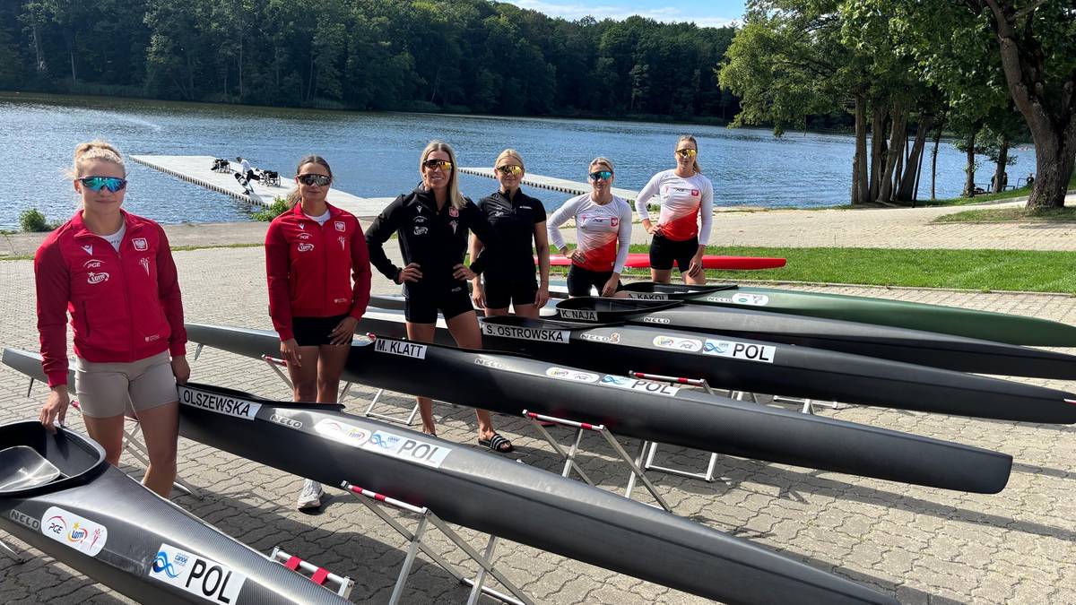 Siedem kobiet w strojach sportowych pozuje do zdjęcia na tle jeziora i kajaków. Wszystkie ubrania reprezentują klub sportowy.