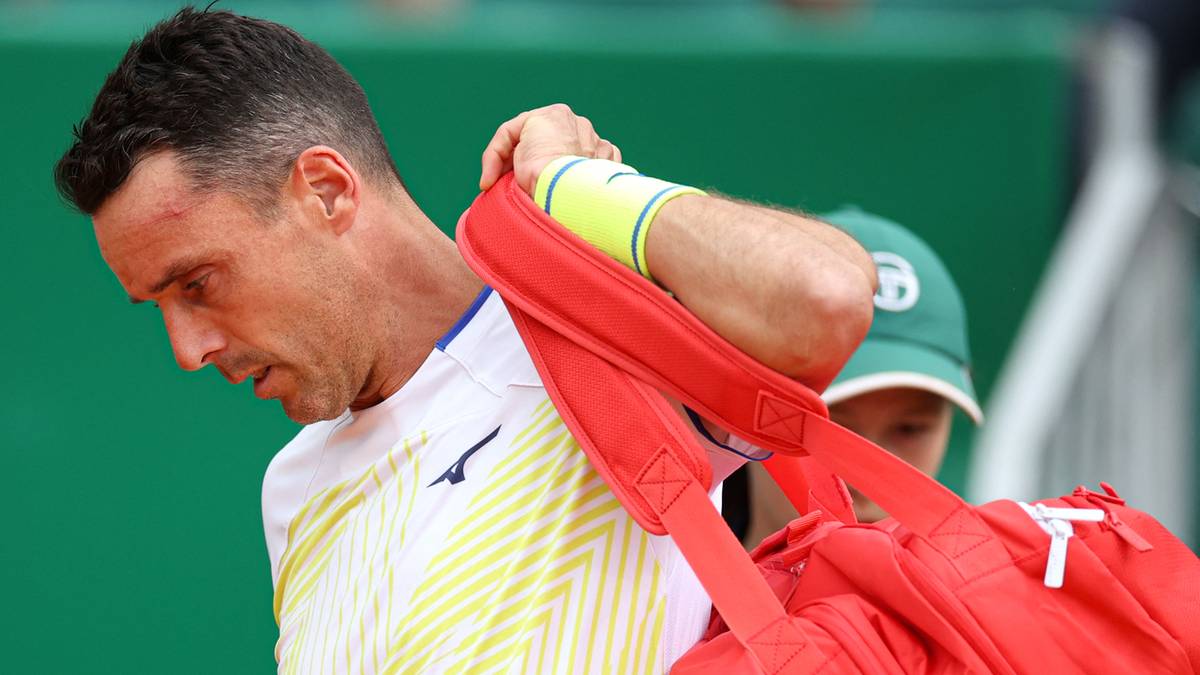 Tenisista z czerwoną torbą sportową na ramieniu, w białej koszulce z żółtymi pasami.