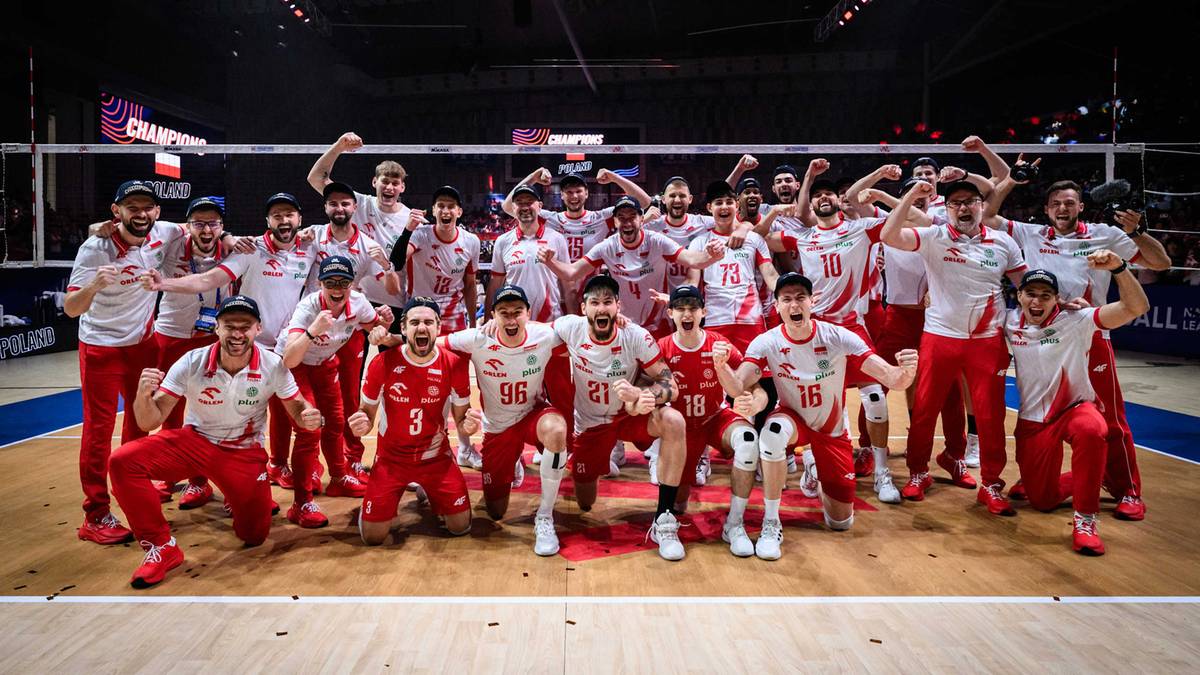 Drużyna siatkarzy w czerwonych koszulkach stoi i siedzi na parkiecie, pozując do zdjęcia po meczu. Większość z nich uśmiecha się i unosi ręce w geście triumfu.