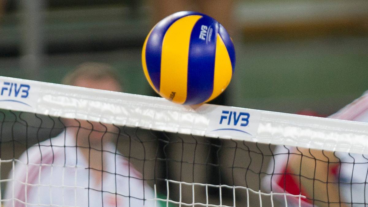 Żółto-niebieska piłka do siatkówki nad siatką z napisem FIVB, z niewyraźnymi sylwetkami zawodników w tle.