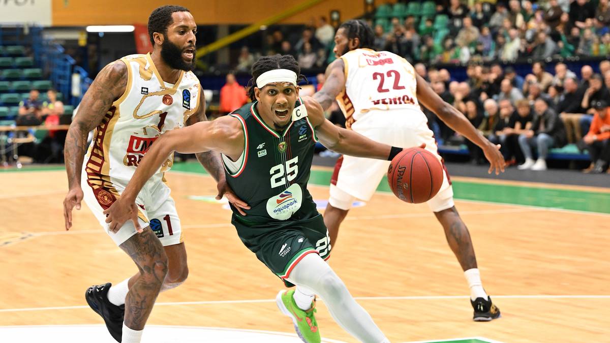 Trzech koszykarzy w akcji na boisku. Gracz w zielonej koszulce z numerem 25 drybluje piłką, mijając dwóch rywali w białych strojach.