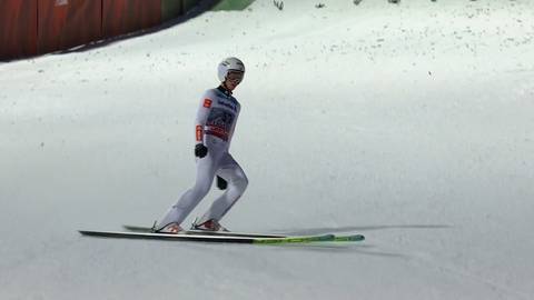 Skoczek narciarski w białym kombinezonie i kasku stoi na nartach na śniegu.