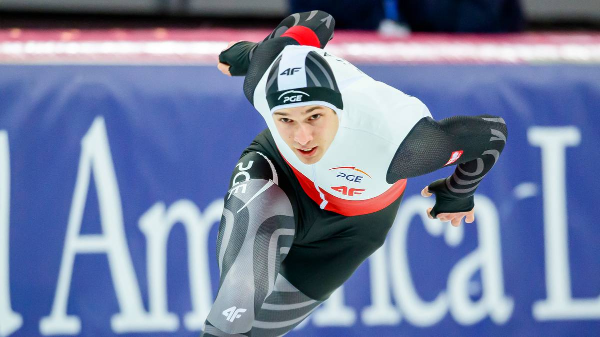 Łyżwiarz szybki w ruchu na lodowisku, w białej koszulce i czarnych spodenkach z szarymi wzorami.