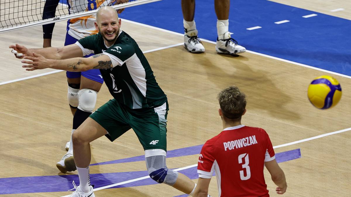 Gracz w zielonej koszulce wykonuje przyjęcie piłki do siatkówki, z zawodnikiem w czerwonej koszulce na pierwszym planie, patrzącym w jego stronę.