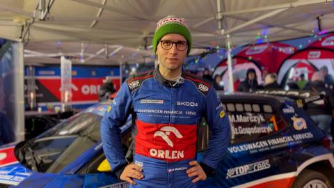 Mikołaj Marczyk, kierowca rajdowy w niebieskim kombinezonie z logo ORLEN i Volkswagen Financial Services, stoi przed samochodem rajdowym.