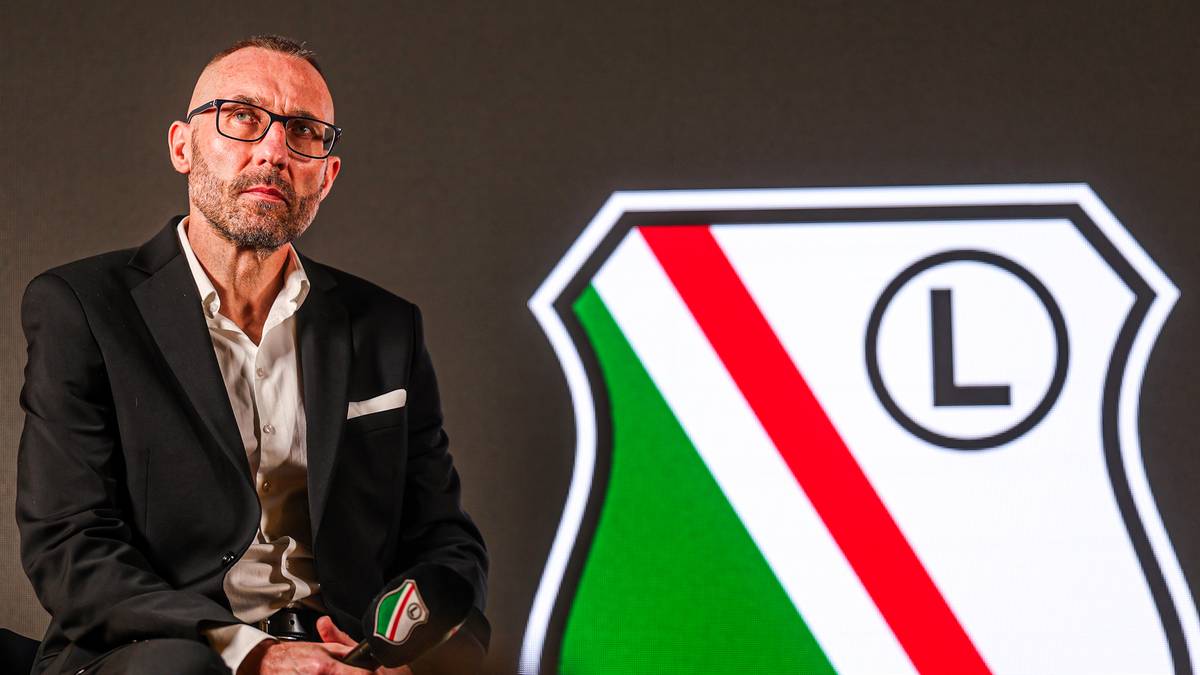 Mężczyzna w okularach siedzi obok herbu Legii Warszawa.