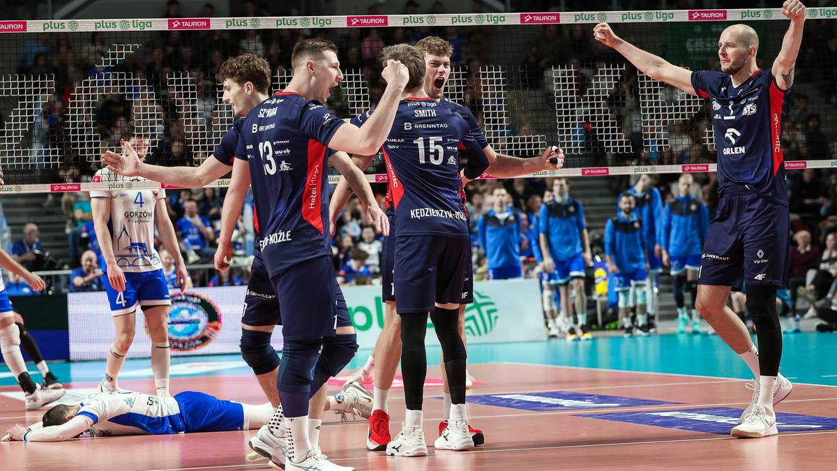Grupa siatkarzy w niebieskich strojach świętuje zwycięstwo na parkiecie, unosząc ręce w geście radości. Jeden z nich klęczy na ziemi.