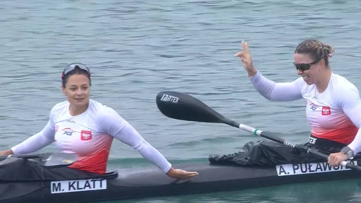 Dwie kobiety w kajaku, jedna z nich podnosi rękę w geście zwycięstwa. Obie ubrane są w stroje sportowe z polskimi symbolami.