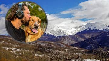 Mężczyzna w słuchawkach całuje psa w policzek na tle górskiego krajobrazu.