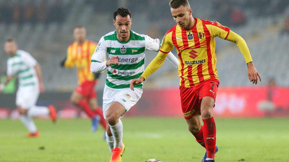 Dw&oacute;ch piłkarzy ścigających się po boisku z piłką, jeden w zielono-białej koszulce, drugi w ż&oacute;łto-czerwonej.