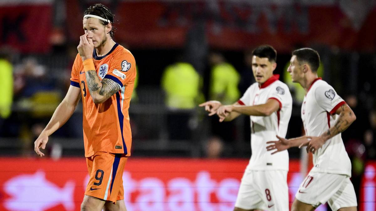 Trzech piłkarzy w białych i pomarańczowych strojach na boisku piłkarskim podczas meczu eliminacyjnego.