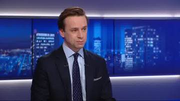 Mężczyzna w garniturze i krawacie, siedzi na tle ekran&oacute;w wyświetlających nocny widok miasta.