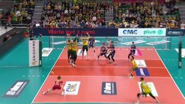 Gracze drużyny Aluron CMC Warta Zawiercie w ż&oacute;łtych strojach podczas akcji meczowej z ZAKSA Kędzierzyn-Koźle. Jeden z zawodnik&oacute;w wykonuje wyskok do ataku, podczas gdy koledzy z drużyny wspierają go na boisku. Widoczni są r&oacute;wnież przeciwnicy i kibice na trybunach.