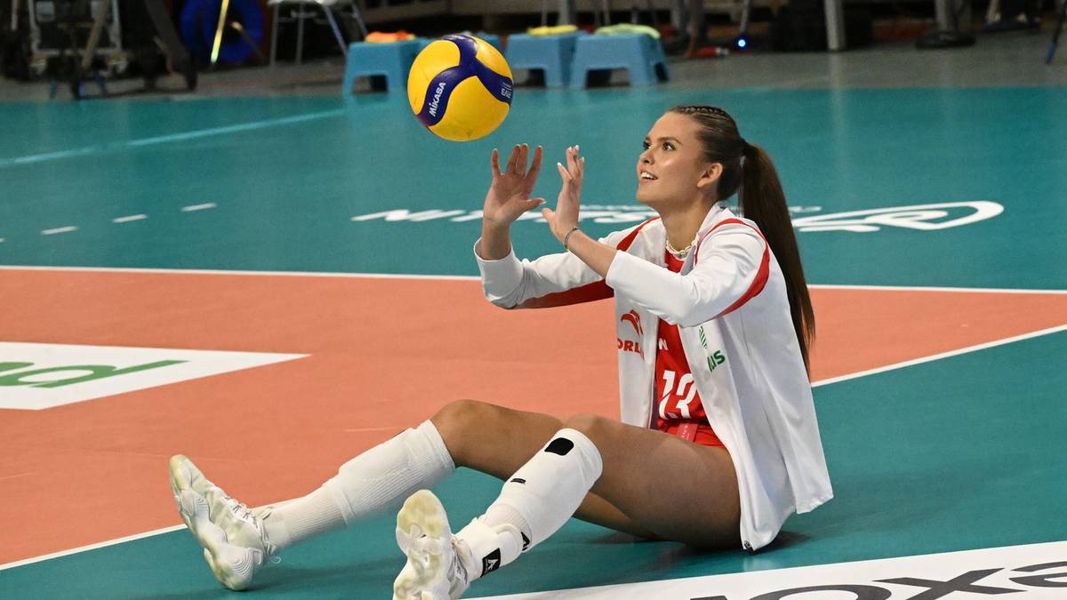 Siatkarka siedząca na parkiecie, łapie piłkę do siatkówki nad głową.