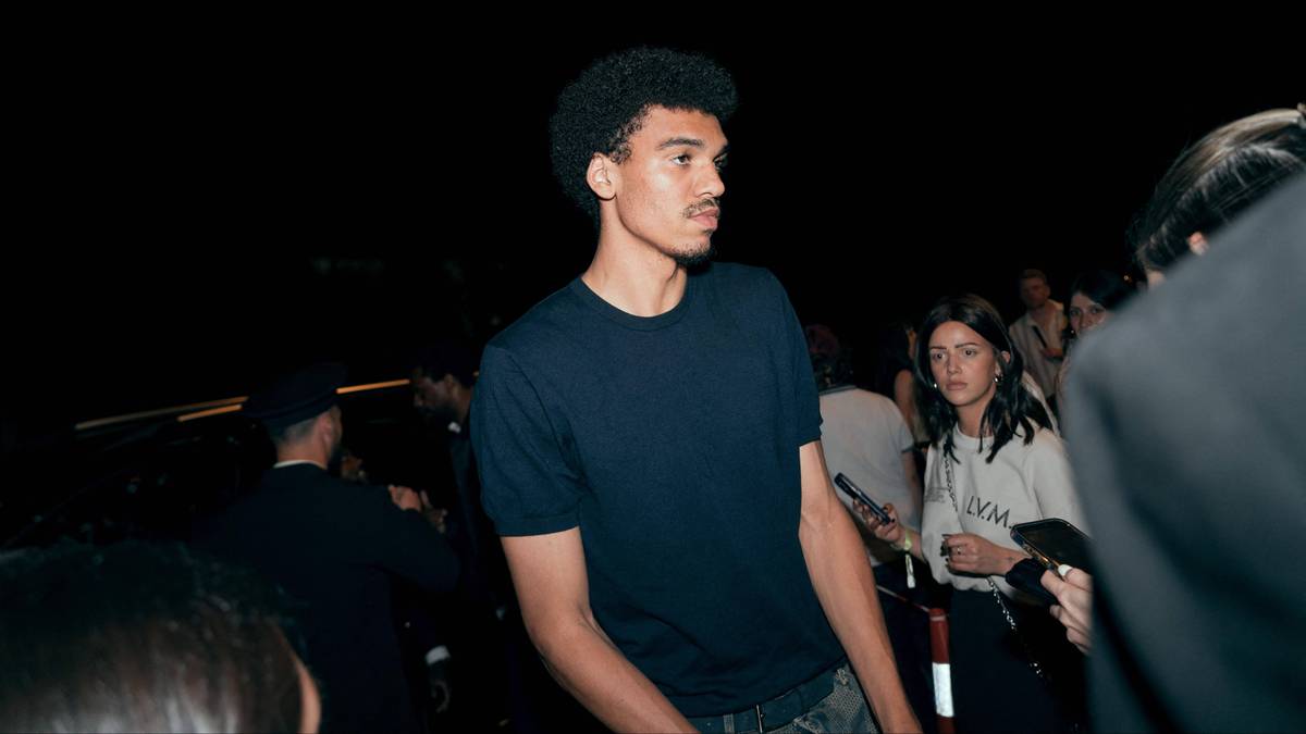 Młody, wysoki mężczyzna o ciemnej karnacji, ubrany w ciemną koszulkę, stoi wśród tłumu ludzi. Ma kręcone, ciemne włosy.