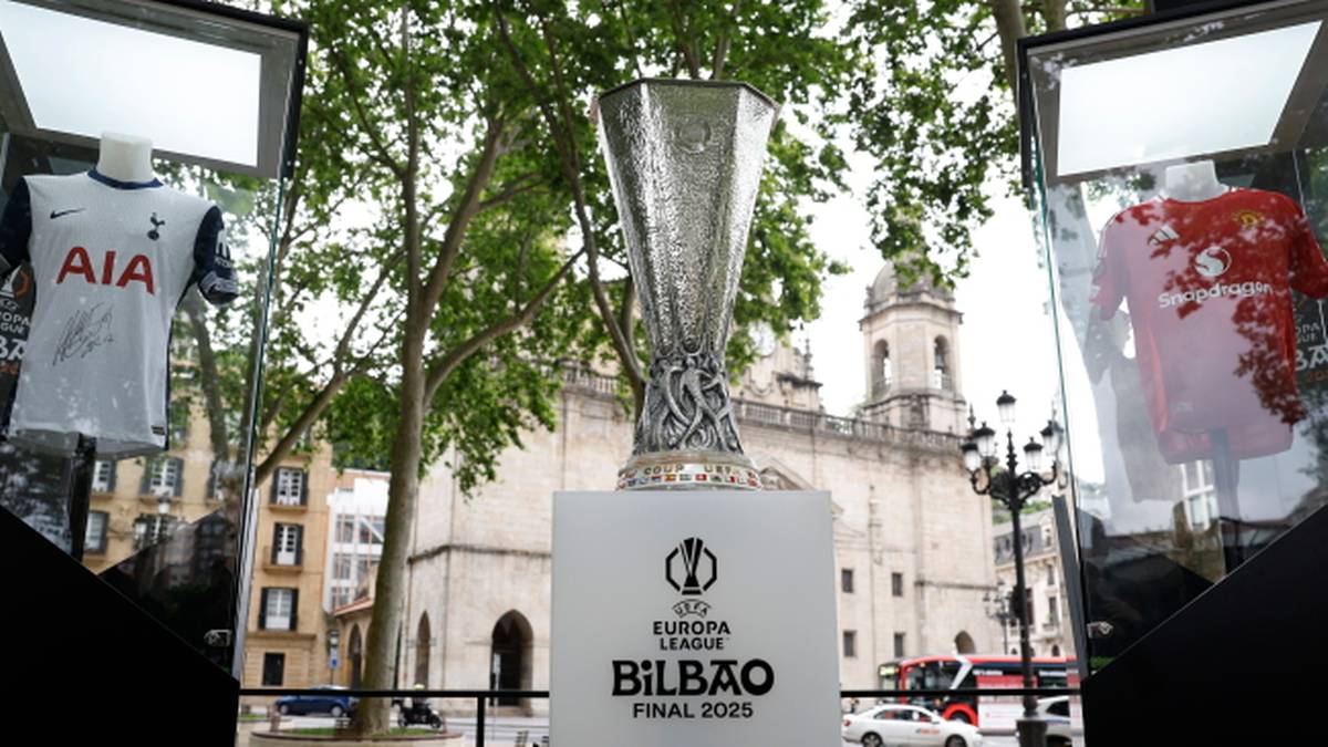 Trofeum Ligi Europy na postumencie z logo i nazwą miasta Bilbao. Po bokach widoczne są wystawione koszulki piłkarskie, po lewej koszulka Tottenhamu, po prawej koszulka w różowym kolorze. W tle budynek z wieżą oraz drzewa i uliczne oświetlenie.