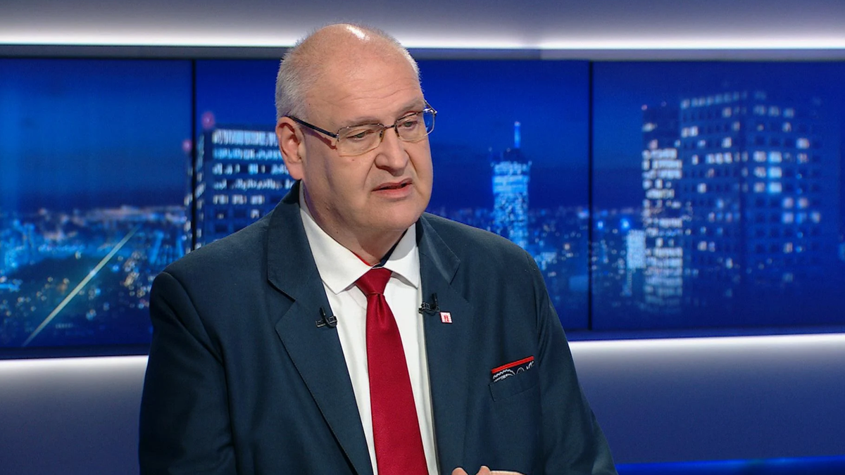 Bogdan Święczkowski, prezes Trybunału Konstytucyjnego, w garniturze i z czerwoną muszką, wypowiada się na tle ekran&oacute;w z nocną panoramą miasta.