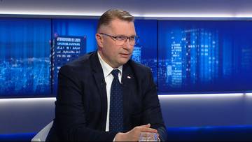 Przemysław Czarnek w garniturze i okularach siedzi w studiu telewizyjnym, na tle ekran&oacute;w wyświetlających nocny widok miasta.