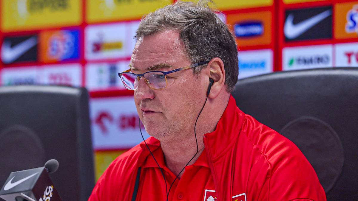 Mężczyzna w okularach i czerwonej koszulce z słuchawkami w uszach, siedzący za stołem z mikrofonem.