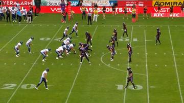 Gracze futbolu amerykańskiego w strojach fioletowo-czarnych i białych na zielonym boisku podczas meczu.