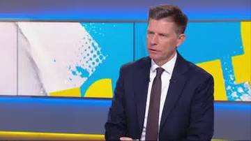 Mężczyzna w garniturze i krawacie siedzi, patrząc w bok z lekko zmarszczonymi brwiami. Tło stanowi jasny, abstrakcyjny wz&oacute;r na niebieskim i ż&oacute;łtym tle.