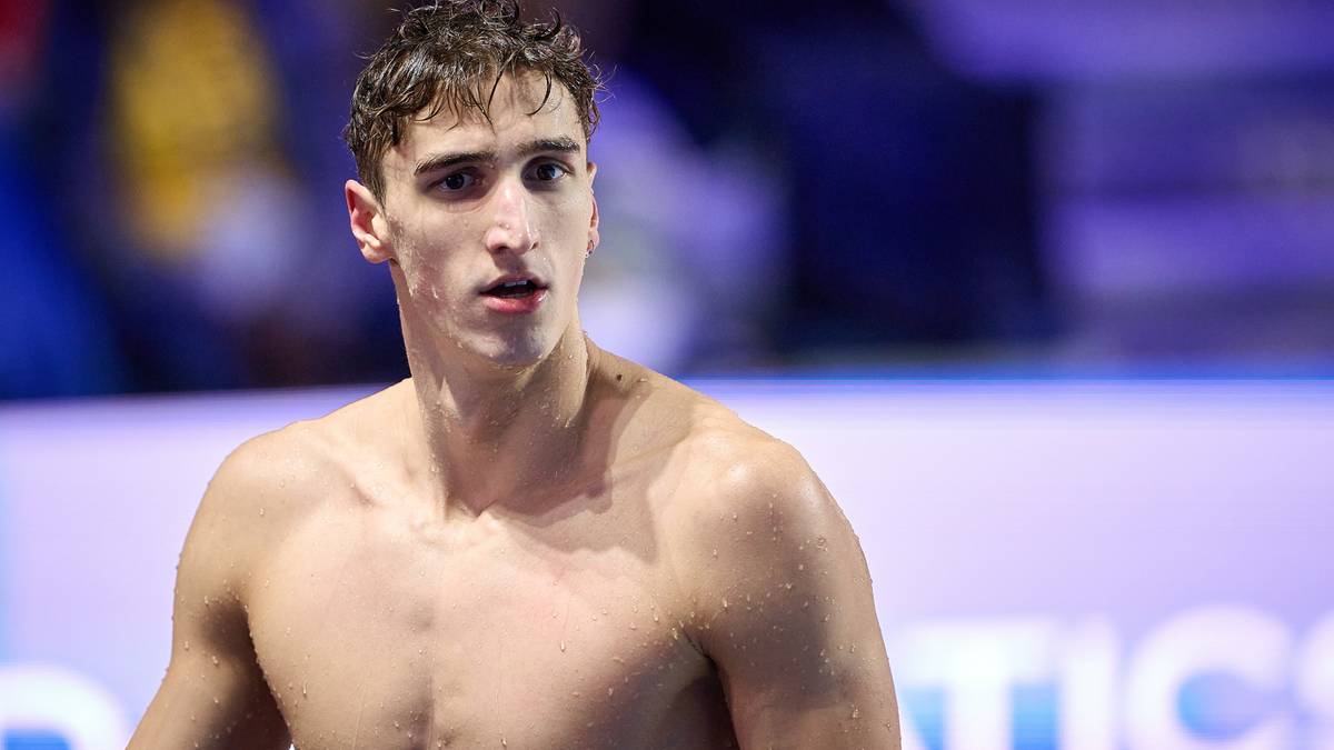 Młody mężczyzna o ciemnych włosach i inteligentnym spojrzeniu, z mokrą skórą i włosami, ubrany tylko w kąpielówki, patrzy w przód z determinacją.