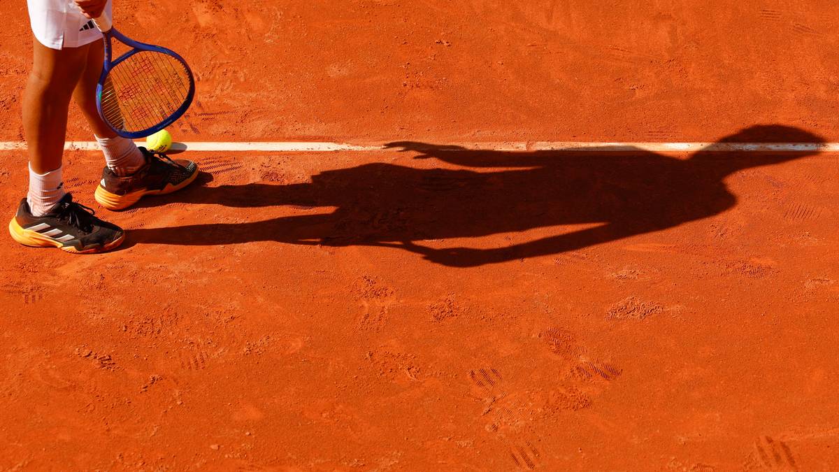 Widok z perspektywy kortu ziemnego na nogi tenisisty z rakietą i jego cień.