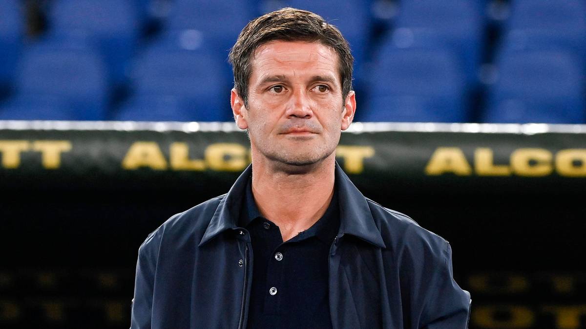 Mężczyzna w średnim wieku, o ciemnych włosach i ciemnych oczach, ubrany w granatową kurtkę i czarny polo, stoi na tle stadionu.