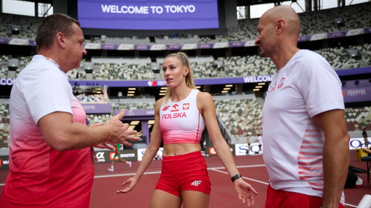Trzy osoby stoją na stadionie lekkoatletycznym. Kobieta w stroju sportowym z napisem "POLSKA" i dwaj mężczyźni, prawdopodobnie trenerzy, prowadzą rozmowę.