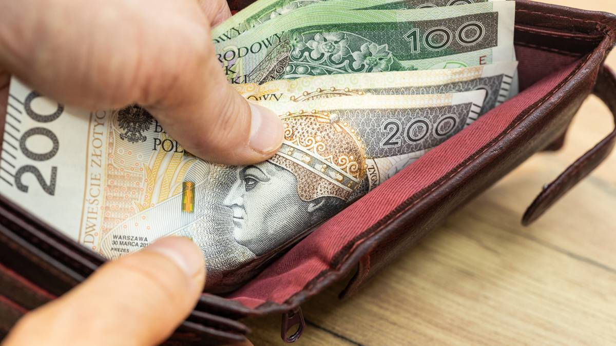 Dłoń umieszcza banknoty o wartości 100 i 200 złotych w brązowym portfelu.