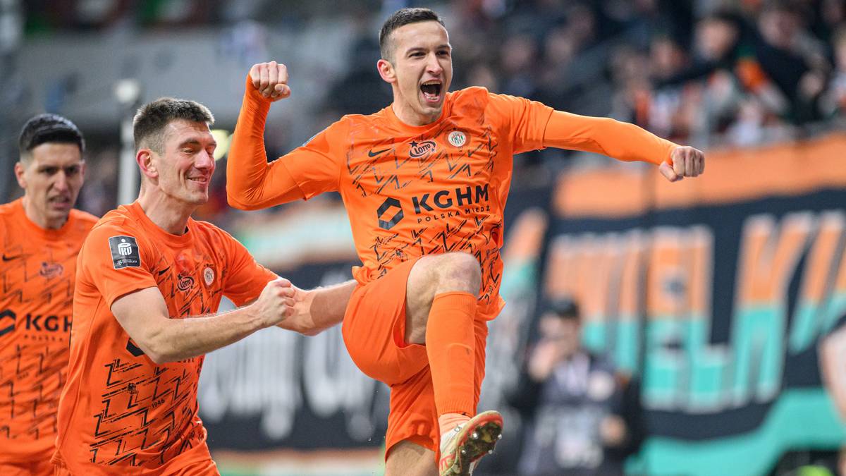 Piłkarz KGHM Zagłębia Lubin, Mateusz Grzybek, cieszy się z zdobytej bramki, z uniesioną ręką i otwartymi ustami, podczas gdy inny zawodnik, Damian Dąbrowski, gratuluje mu.