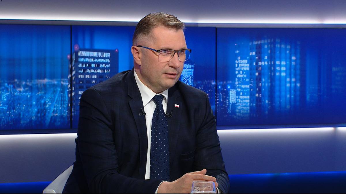 Przemysław Czarnek w garniturze i okularach siedzi w studiu telewizyjnym, na tle ekran&oacute;w wyświetlających nocny widok miasta.
